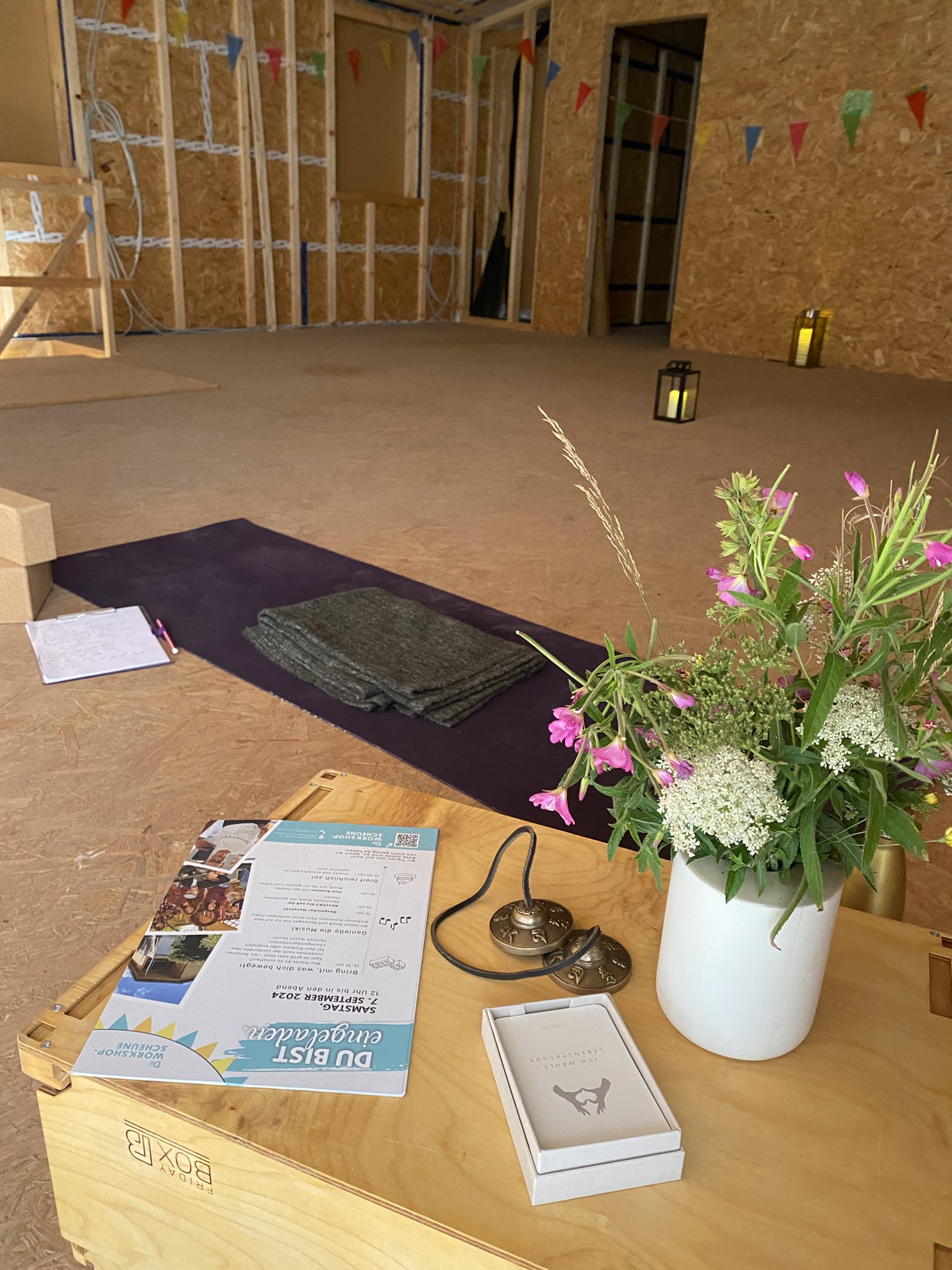 Yoga-Setup im Rohbau. Wir sehen eine Yogamatte, Wiesenblümchen in einer Vase, Zimbeln und Karten.
